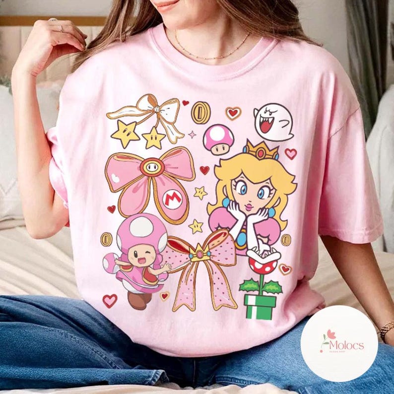 Puede incluir: Camiseta rosa claro con un gr&aacute;fico colorido de la Princesa Peach, un hongo, estrellas, lazos y otros personajes de Super Mario Bros. El dise&ntilde;o incluye corazones y monedas. La camiseta tiene un corte holgado.