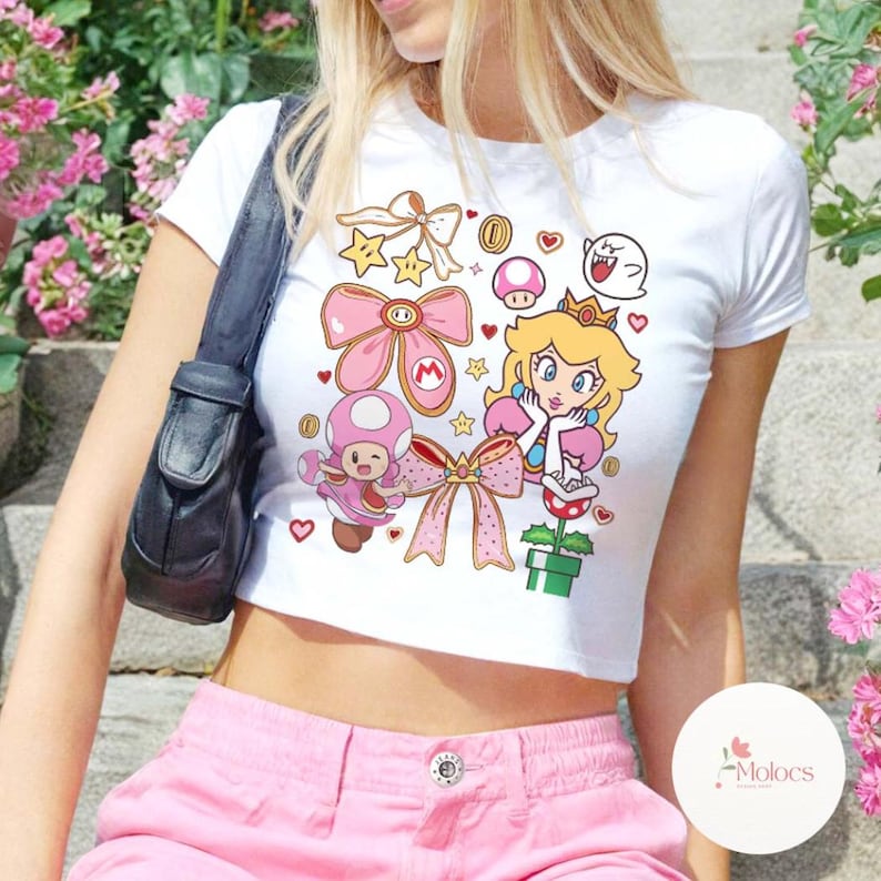 Puede incluir: Top corto blanco con un gr&aacute;fico colorido de la Princesa Peach, Toadette y otros personajes de Super Mario Bros. El dise&ntilde;o incluye lazos rosas, estrellas, monedas y corazones. La camiseta tiene cuello redondo y mangas cortas.