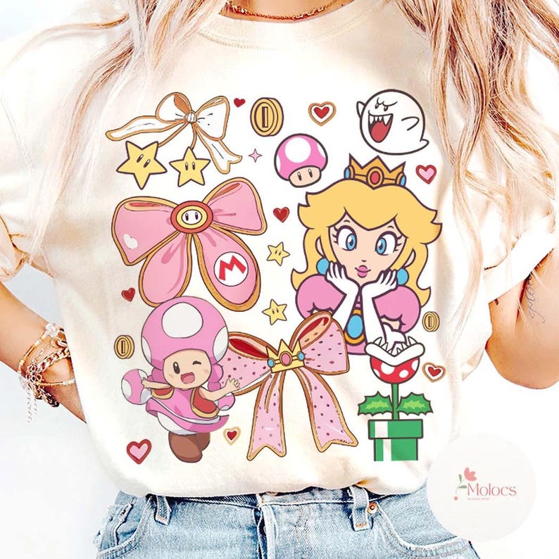 Puede incluir: Camiseta de color crema con un dise&ntilde;o gr&aacute;fico colorido con personajes y elementos de la serie de videojuegos Super Mario. El dise&ntilde;o incluye a la Princesa Peach, Toad, monedas, estrellas y lazos.