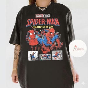 Op de afbeelding: Zwart T-shirt met een Spider-Man-ontwerp van Marvel Studios. Het ontwerp bevat de tekst "Spider-Man Brand New Day" in rood en blauw, samen met stripboekillustraties van Spider-Man in actie.