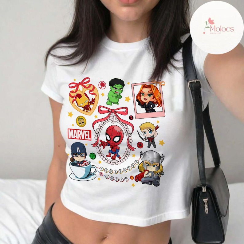 May include: White crop top with a colorful graphic print of various Marvel characters, including Spider-Man, Iron Man, Hulk, Black Widow, Captain America, and Thor. The graphic includes the word "MARVEL" and a red ribbon with a bow.