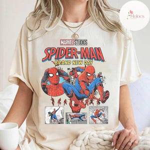 Op de afbeelding: Cr&egrave;mekleurig T-shirt met een afbeelding van Spider-Man in actieposes. Het ontwerp bevat de tekst "MARVEL STUDIOS SPIDER-MAN BRAND NEW DAY" in rood, blauw en geel. Het shirt heeft korte mouwen en een ronde hals.