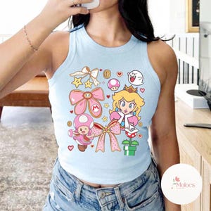 Puede incluir: Camiseta sin mangas azul claro con un gr&aacute;fico colorido de la Princesa Peach, Toad y otros personajes de la serie de videojuegos Super Mario. El dise&ntilde;o incluye lazos rosas, estrellas, monedas y corazones. La camiseta sin mangas es sin mangas y tiene cuello redondo.
