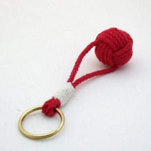 May include: A red rope keychain with a white accent and a brass key ring. The rope is tied into a knot at the top.