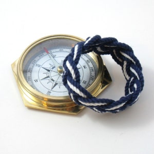 May include: A gold compass with a white face and black markings. The compass is surrounded by a blue and white rope bracelet.