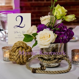 May include: A table number card with the number "2" and the text "Jo Ann & Matt Tied the Knot" is displayed on a table with a centerpiece of white and purple flowers in a silver vase. A thick rope is wrapped around the vase and a large rope knot is on the table.