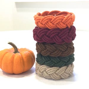 May include: A stack of five braided rope bracelets in fall colors. The bracelets are stacked from top to bottom: orange, burgundy, brown, green, and beige.