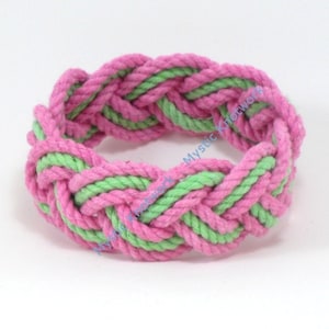 May include: A pink and green braided rope bracelet. The rope is woven in a pattern that creates a series of knots.