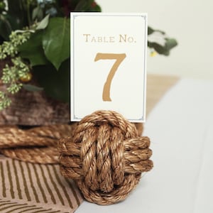 May include: A table number card with the text "Table No. 7" in gold lettering, held by a decorative rope knot. The knot is made of thick, brown rope. The card has a decorative border.