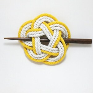 May include: A yellow and white rope knot hair pin. The knot is tied in a decorative pattern and is attached to a wooden stick.