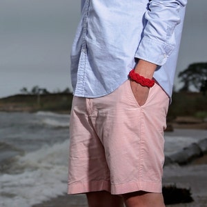 May include: A man wearing a light blue button-down shirt and pink shorts. He is wearing a red braided bracelet on his wrist.