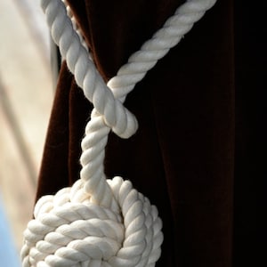 May include: White rope tied into a knot, used as a curtain tieback. The rope is looped around a brown curtain and tied into a large knot at the bottom.