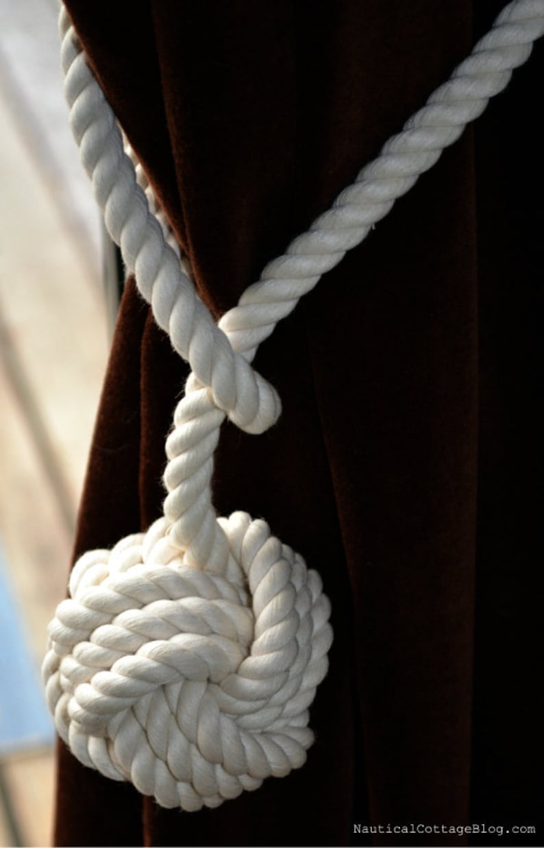 May include: White rope tied into a knot, used as a curtain tieback. The rope is looped around a brown curtain and tied into a large knot at the bottom.