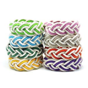May include: A collection of eight circular rope coasters arranged in two stacks. Each coaster features a braided design combining white rope with a secondary color, including lime green, light blue, dark green, yellow, pink, beige, orange, and purple.