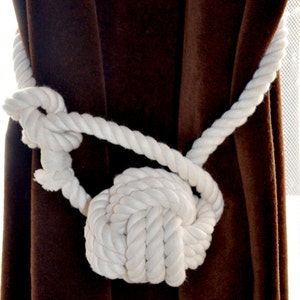 May include: White rope tied in a knot to create a decorative curtain tieback. The tieback is against a brown velvet curtain.