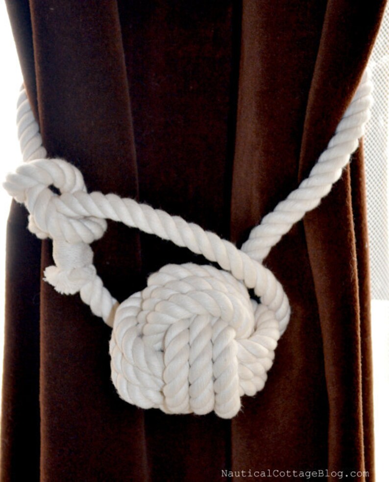 May include: White rope tied in a knot to create a decorative curtain tieback. The tieback is against a brown velvet curtain.
