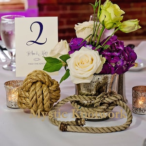 May include: A wedding table centerpiece with a white and purple flower arrangement in a silver vase. The table number is 2, and the text "Jo Ann & Matt Tied the Knot" is on the card. A large rope knot is on the table, and the text "Mystic Knotwork" is on the table cloth.