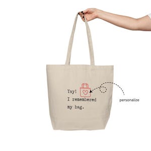 May include: A beige tote bag with a red heart and the text "Yay! I remembered my bag." The bag has a small red bag with a heart on it in the top left corner. The text "personalize" is written in black next to a dashed line that circles the small red bag.