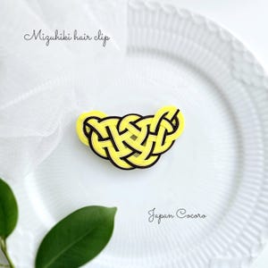 以下が含まれることがあります： 白い背景に「Mizuhiki hair clip」と「Japan Cocoro」の文字が書かれた、黄色と黒の編組紐製のヘアクリップです。