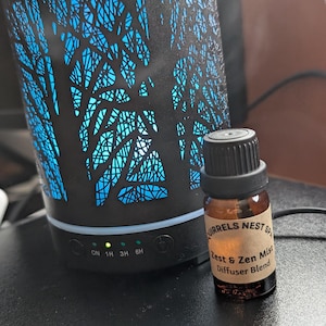 May include: A black diffuser with a tree-like design and blue light. The diffuser has a timer with settings for 1, 3, and 6 hours. A small brown bottle of essential oil is in front of the diffuser. The bottle label reads "Squirrels Nest Spa Zest & Zen Mist Diffuser Blend".