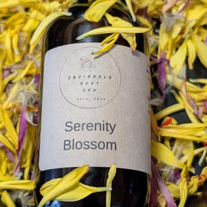 May include: A dark brown bottle with a black cap, labelled "Serenity Blossom" from Squirrels Nest Spa, is surrounded by yellow and purple flower petals. The label has a squirrel logo and the text "ESTD 2020".