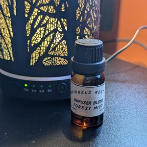 May include: A small brown glass bottle with a black cap and a white label that reads "Squirrels Nest Diffuser Blend Forest Mist".