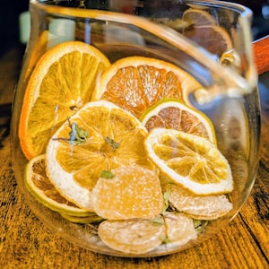 May include: A glass jug filled with dried citrus fruit slices, including orange, lemon, and lime, and pieces of candied ginger. The jug is sitting on a wooden surface.