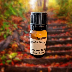 May include: An amber glass bottle with a black cap, labelled "Autumn Aura Diffuser Blend." The bottle is set against a blurred background of a forest path with autumn leaves. The product is from "Squirrels Nest Spa."