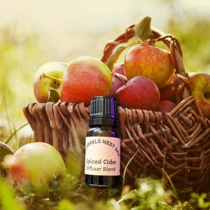 May include: A bottle of Spiced Cider Diffuser Blend is positioned in front of a wicker basket filled with red and green apples. The bottle has a black cap and a label with the text "Squirrels Nest Spa". The scene is outdoors.