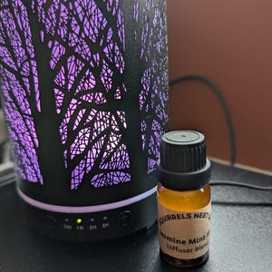 May include: A black essential oil diffuser with a tree-like design and a purple light. The diffuser has buttons labelled "ON", "1H", "3H", and "6H". A small amber glass bottle of essential oil is in front of the diffuser. The bottle has a black cap and a label that reads "Squirrels Nest Jasmine Mint Diffuser Blend".
