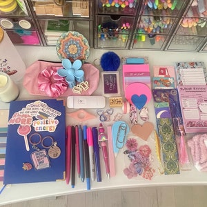 May include: A flat lay of various stationery and craft supplies, including a blue notebook with colorful stickers, pink and blue hair scrunchies, a pink fuzzy slipper, a set of colorful pens, and a pink and blue notepad with the text "Book Review".