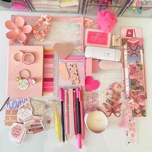 May include: A flat lay of pink and white stationery supplies, including pens, pencils, stickers, washi tape, and a small candle. The items are arranged on a white surface and include the text "Make yourself a priority".