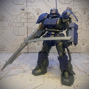 以下が含まれることがあります： 濃い紫と灰色のロボットのアクションフィギュアで、大きな銃と剣を持っています。フィギュアは白い背景に立っています。