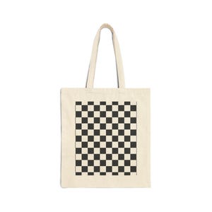 May include: A rectangular off-white canvas tote bag featuring two long shoulder straps and a centered black and white checkered pattern. The design consists of a grid of alternating dark and light squares printed on the front of the fabric.