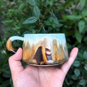 Peut inclure: Une tasse en céramique faite à la main avec un design unique. La tasse présente un bord bleu clair qui se transforme en tons bruns et noirs. La poignée est bleu clair avec un accent marron. La tasse est tenue dans une main sur un fond de feuilles vertes.