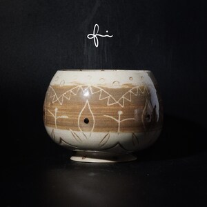 Peut inclure: Un bol en céramique fait à la main, de couleur marron et crème. Le bol présente un motif décoratif de formes géométriques et de figures stylisées. Le bol est posé sur un fond sombre, soulignant son design et sa texture uniques.