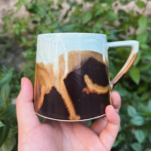 Può includere: Una tazza in ceramica fatta a mano con una smaltatura unica. La tazza presenta una sfumatura di colori, tra cui azzurro, marrone e marrone scuro. Il manico ha un design semplice e angolare. La tazza è tenuta in una mano, con uno sfondo di foglie verdi.