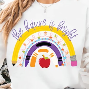 May include: A white crewneck sweatshirt featuring a graphic of a rainbow made from pencils and crayons with the text "the future is bright" in purple script. A red apple is centered at the base of the rainbow, surrounded by small orange and blue stars.