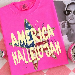 Pu&ograve; includere: Una maglietta girocollo rosa acceso con le parole "AMERICA HALLELUJAH" in giallo sbiadito. La maglietta presenta un design a stella con i colori della bandiera americana e dettagli dorati. La maglietta &egrave; piegata su una superficie a scacchi rossi e bianchi.
