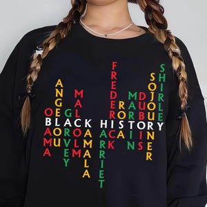 May include: Black sweatshirt with the words "BLACK HISTORY" in white, and names of historical figures in red, green, and yellow. The sweatshirt has long sleeves and a crew neck.