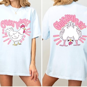 May include: Light blue t-shirt with a cartoon chicken design. The front of the shirt says "Guess What?" above a white chicken with pink accents. The back of the shirt says "Chicken Butt" with a chicken graphic.