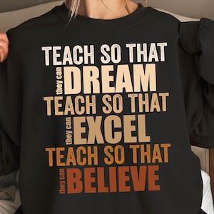 May include: Black sweatshirt with white and tan text. The text reads "TEACH SO THAT DREAM, TEACH SO THAT EXCEL, TEACH SO THAT BELIEVE." The words "they can" are written vertically on the left side of the text.