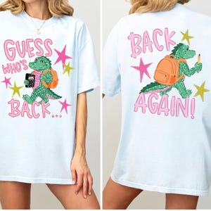 以下が含まれることがあります： 緑色のワニのグラフィックが描かれた水色のTシャツ。前面には「GUESS WHO'S BACK...」という文字があり、ワニが黒いノートとオレンジ色のバックパックを持っています。背面には「BACK AGAIN!」という文字と、鉛筆を持ったワニが描かれています。