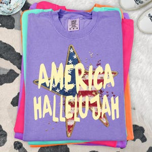 Pu&ograve; includere: Una pila di magliette colorate, con una maglietta viola sopra. La maglietta viola presenta un disegno a stella con la bandiera americana e le parole "AMERICA HALLELUJAH" in giallo. Le magliette sono impilate su una superficie a motivi.
