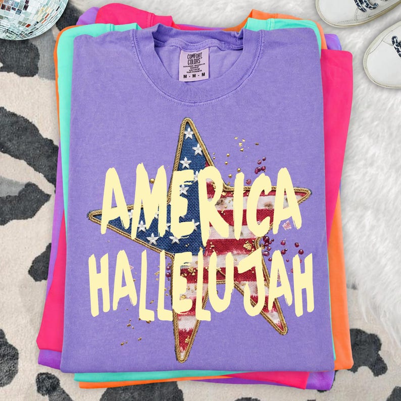 Pu&ograve; includere: Una pila di magliette colorate, con una maglietta viola sopra. La maglietta viola presenta un disegno a stella con la bandiera americana e le parole "AMERICA HALLELUJAH" in giallo. Le magliette sono impilate su una superficie a motivi.