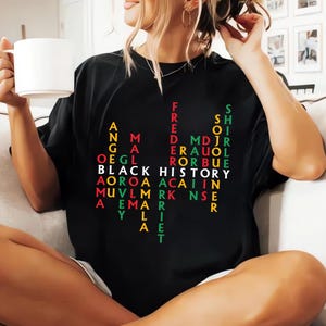 May include: Black t-shirt with the words "BLACK HISTORY" in white, and names of historical figures in various colors. The shirt has a crew neck and short sleeves. The person is holding a white mug.