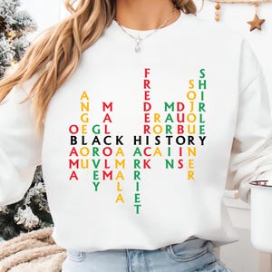 May include: White sweatshirt with the words "BLACK HISTORY" in black, and other names in various colors. The names are arranged vertically and horizontally. The sweatshirt is a crew neck style.