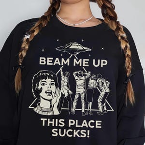May include: Black crew neck sweatshirt with a retro graphic design. The design features a UFO, people looking up, and the phrases "BEAM ME UP" and "THIS PLACE SUCKS!" in white text. The sweatshirt has long sleeves.