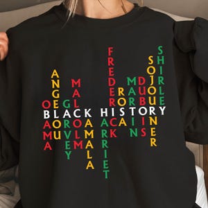 May include: Black sweatshirt with the words "BLACK HISTORY" in white, and other names in yellow, green, and red. The sweatshirt has a crew neck and long sleeves. The text is arranged vertically and horizontally.