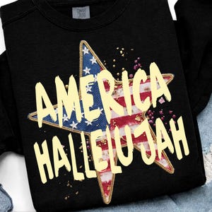 Pu&ograve; includere: Felpa nera con un design a stella con i colori della bandiera americana e le parole "AMERICA HALLELUJAH" in giallo. La stella ha un bordo dorato e macchie dorate.